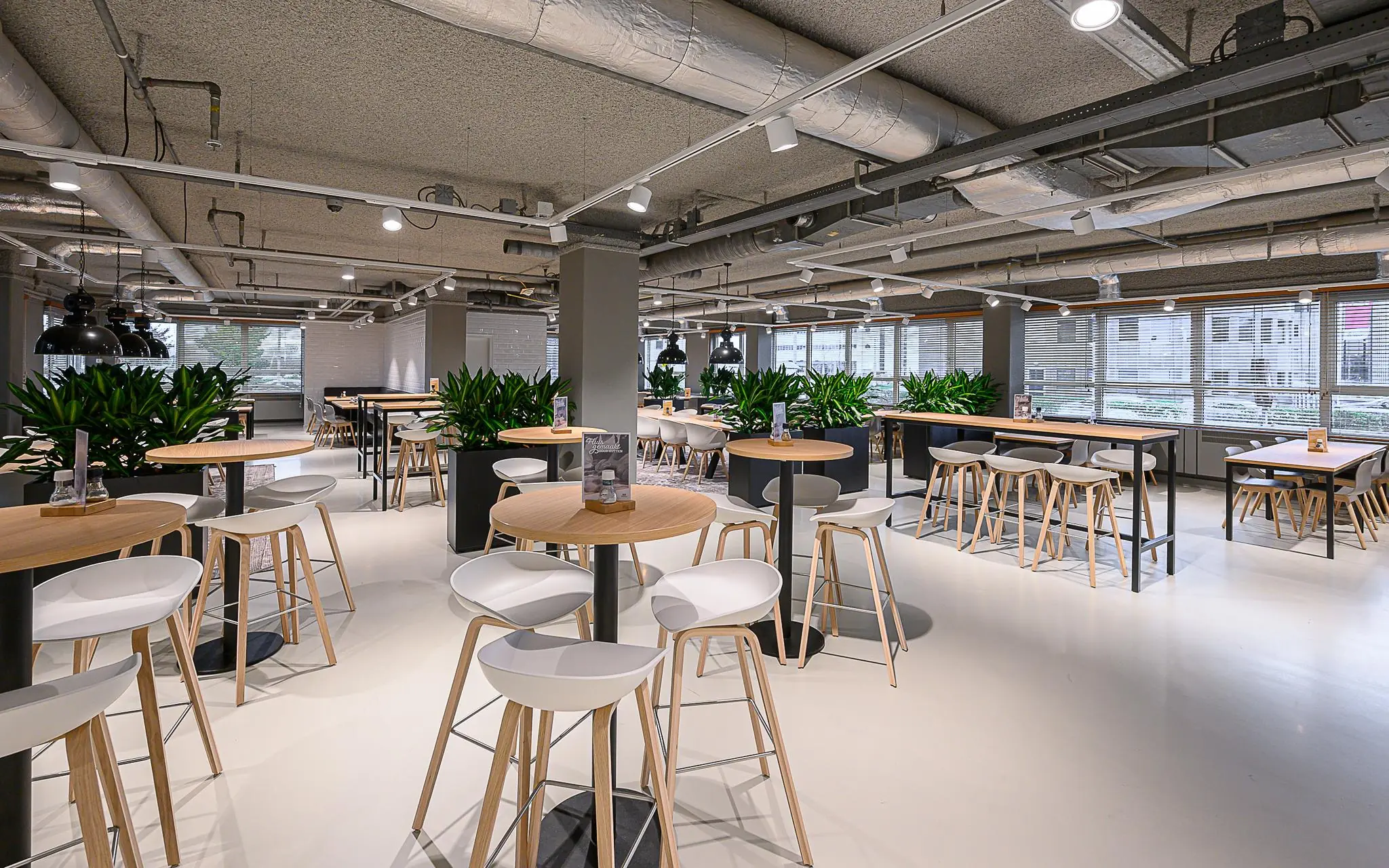 Moderne, open bedrijfsrestaurant in Rivium Quadrant met hoge tafels, barkrukken en groene planten als scheidingselementen.